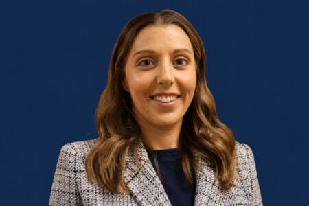 A woman with long wavy brown hair, wearing a patterned blazer, smiles directly at the viewer against a plain dark blue background.