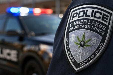 Finger Lakes Drug Task Force police officer's patch, featuring a cannabis leaf, with a flashing patrol car behind.