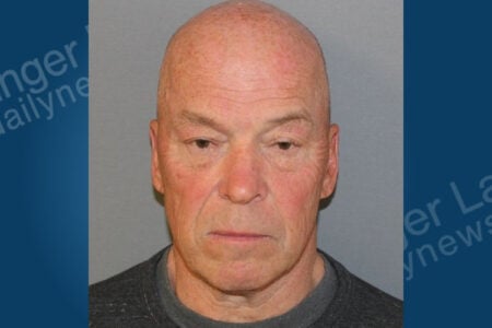 Mugshot of a bald older man with a neutral expression, wearing a dark shirt against a plain grey background.