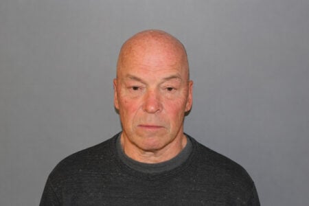 Bald man with a serious, flushed face in a dark grey shirt against a plain grey background.