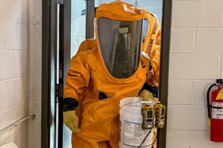 HAZMAT responder in orange suit walks through a doorway, carrying a bucket with monitors, in a cinder block hallway.