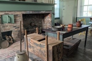 An antique butter churn and primitive washing machine in a historical room with a large brick fireplace.