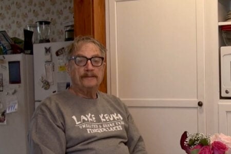 Man with glasses and mustache wearing a "Lake Keuka" sweatshirt, seated in a kitchen.