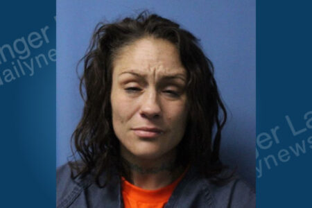 Woman with dark hair, furrowed brow, and neck tattoos in a mugshot, wearing an orange shirt and blue uniform against a blue background.