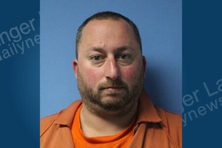 A man with short brown hair and a beard, wearing an orange uniform, stares directly at the camera against a blue background.
