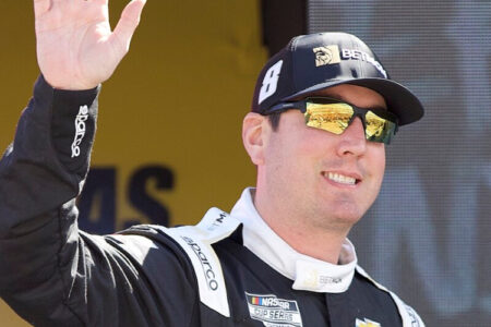 NASCAR driver Kyle Busch smiles and waves in a black racing suit and reflective sunglasses.