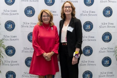 Woman in red dress and woman in black blazer smile in front of a New York State Senate branded backdrop.