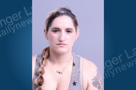 Woman with braided hair, nose ring, "MOM" necklace, and arm tattoo looks directly at the camera for a mugshot.