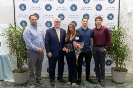 New York State Senator and four others, including an honoree holding a plaque and roses, pose in front of a Senate banner.
