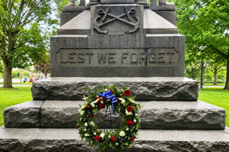 A red, white, and blue wreath sits at the base of a stone war memorial engraved 'LEST WE FORGET' in a town park.