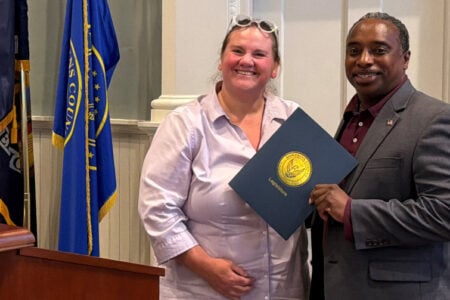 Smiling woman and man holding a Tompkins County Legislature document in a room with flags.