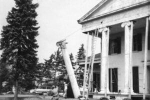 Workers hoist a large fluted Ionic column into position on the front portico of a stately building.