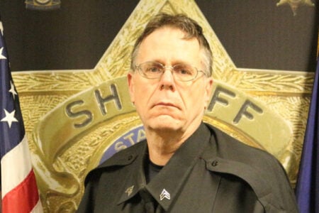 Sheriff's Deputy in black uniform and glasses, standing before a large sheriff's badge and US flag.