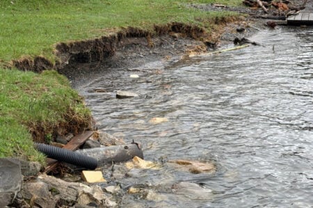 Eroded grassy shoreline of Seneca Lake with a drainage pipe and debris in the water.