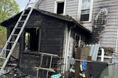 House exterior severely burned by fire, with charred wood siding, melted sections, and a ladder leaning on it.