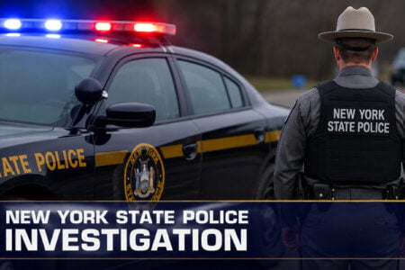 New York State Police officer, back to camera, by patrol car with flashing red and blue lights on a roadside.