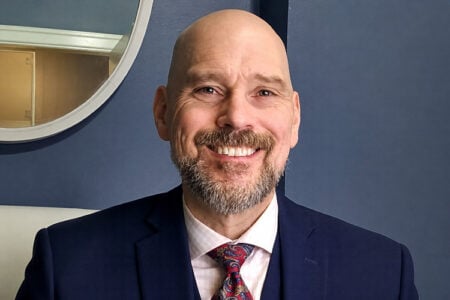 Bald man with a beard in a suit smiles against a blue wall with an oval mirror.