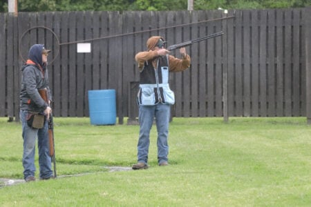 Two men at a Finger Lakes outdoor shooting range: one aims a shotgun, the other observes with his gun, backed by a wooden fence.