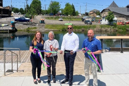 Four people perform a ribbon-cutting ceremony on a waterfront dock next to a canal.