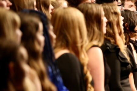 A chorus of students, mostly young women, standing and singing on a dark stage.