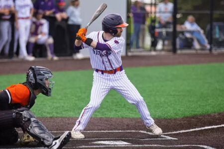 Batter in a white pinstripe, purple & orange uniform stands at home plate with a bat, facing a catcher.