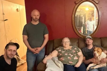 Four adults and a dog pose together in a living room, with a fifth man visible in a mirror reflection.