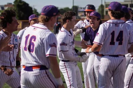 College baseball players in white pinstripes and purple hats celebrate with shouts and embraces on a sunny field.