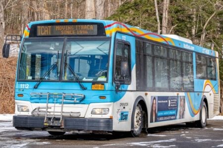 A blue and white TCAT bus with "Moving Ithaca Since 1996" on its display is parked in a snowy wooded area.