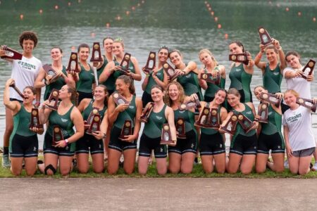 Herons Rowing team poses with numerous NCAA trophies by a lake, celebrating their victory.