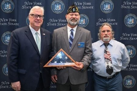 Three men, including veteran Denis Oliver holding his framed flag, stand before a New York State Senate backdrop.