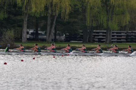 Eight women in a crew shell row on a lake, past trees and RVs on the far shore.