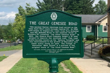 Green historical marker about "The Great Genesee Road" stands on a Marcellus sidewalk, with buildings & street.