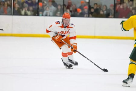 Hobart hockey player in orange and white skates with a stick on the ice during a game, an opponent in yellow and green.