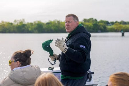 Man in a Herons Rowing jacket speaks from a boat on a lake, holding a green cap and sunglasses.