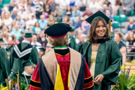 Smiling graduate in green cap and gown looks toward a faculty member during an indoor ceremony, audience visible.