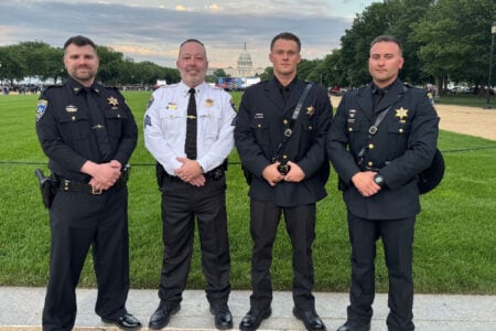 Four uniformed law enforcement officers pose on a grassy lawn with the U.S. Capitol dome in the background.
