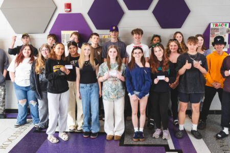 Over two dozen students pose in a school hallway, many holding small white cards.