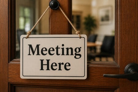 White "Meeting Here" sign hanging on a dark wooden door with a glass panel, revealing a blurred office interior.