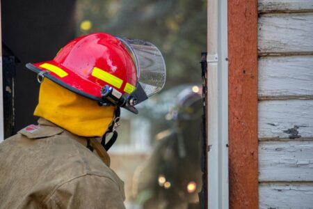 Firefighter in a red helmet and tan coat peers into a dark building opening with peeling paint.