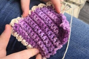 Hands use a crochet hook to add a beige border to a purple yarn swatch, resting on a lap in blue jeans.