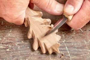 Hands carve a wooden oak leaf shape with a carving knife on a rustic wooden surface.