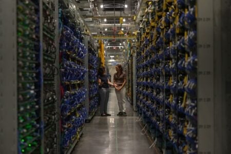 Two women stand talking in a large data center aisle flanked by tall racks of servers with blue and yellow cables.