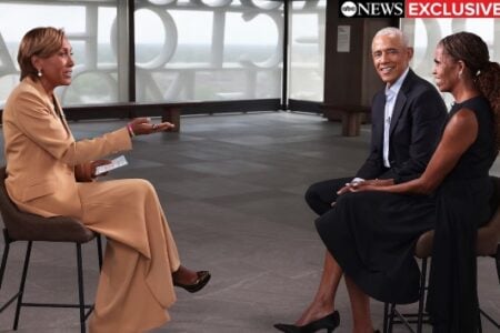 Robin Roberts interviews Barack and Michelle Obama, seated and smiling in a modern room with distinctive windows.