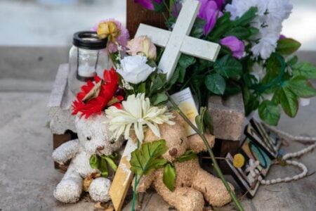 A memorial featuring two teddy bears, a white cross, artificial flowers, and a candle sits on concrete.