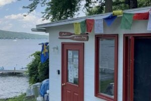 White "Ann's Boathouse Pottery" shop with red door and colorful flags sits by a Finger Lake shoreline.