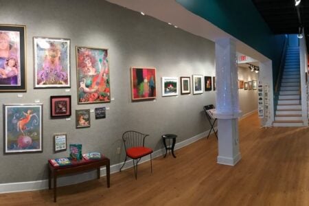 Vibrant artworks adorn the walls of a spacious, modern art gallery, with a staircase and merchandise displays visible.