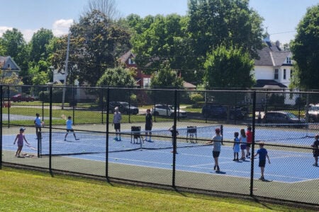 Children and adults on blue tennis courts during a lesson, with houses and trees in the background.