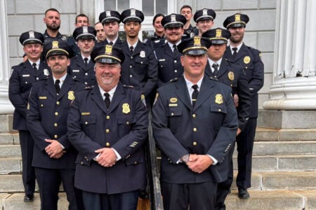 A group of smiling police officers in uniform and caps pose for a photo on stone steps in front of a building with columns.