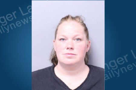 A woman with light hair pulled back and blue eyes poses for a mugshot on a plain background.