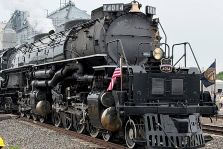 Union Pacific Big Boy #4014 steam locomotive on tracks, emitting smoke, by grain silos with spectators.